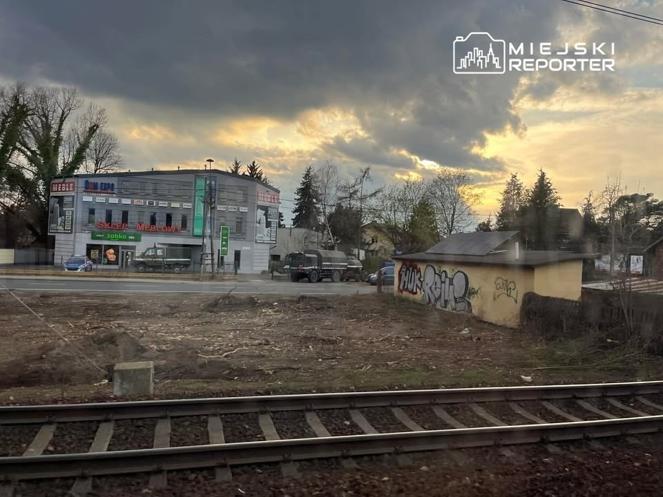 Nowoczesny budynek sklepu meblowego z reklamami, w tle chmurne niebo, a w pierwszym planie tor kolejowy obok wykarczowanego terenu.