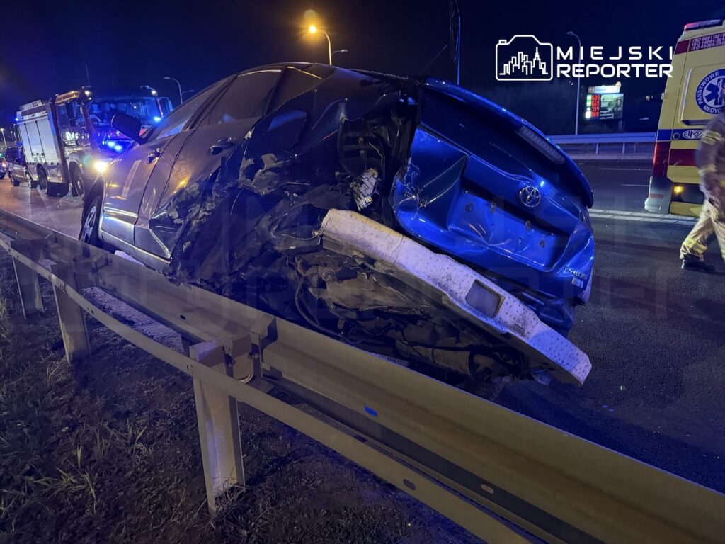 Czarny samochód osobowy z uszkodzoną tylną częścią unosi się na barierze ochronnej przy drodze, w tle widać pojazdy służb ratunkowych.