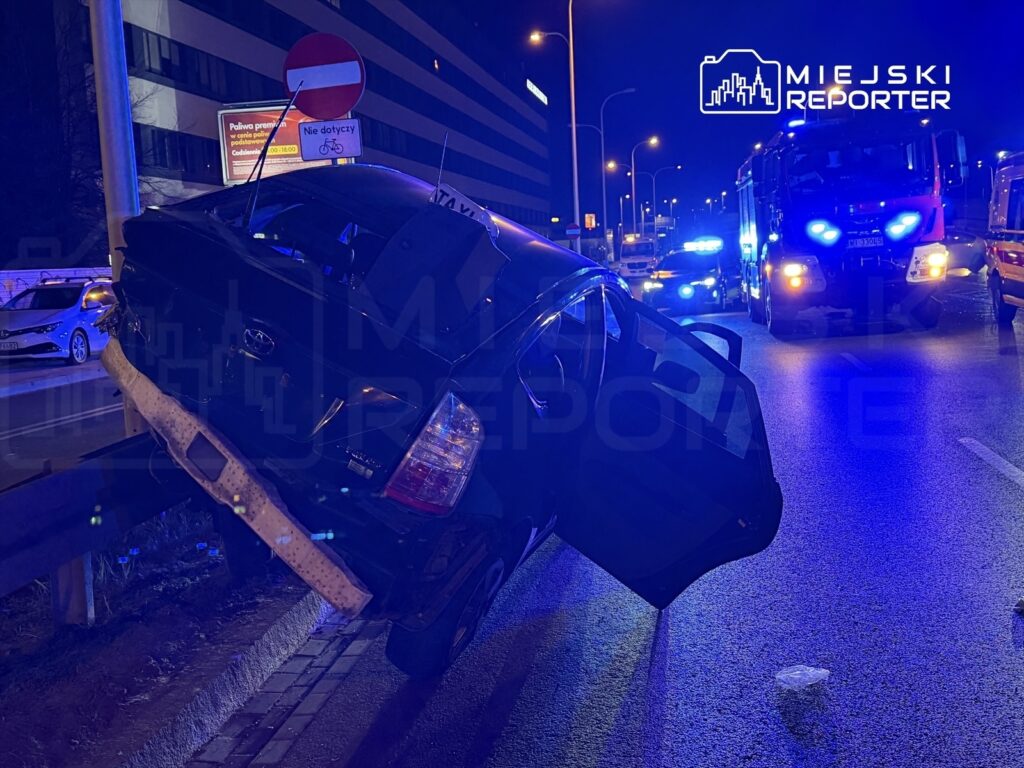 Czarny samochód osobowy unosi się nad krawężnikiem, z otwartymi drzwiami, obok policyjne i strażackie pojazdy na ulicy nocą.