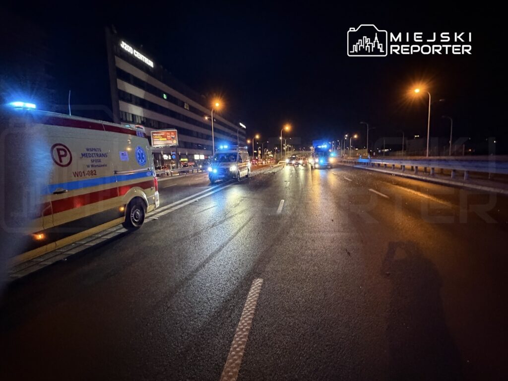 Karetka pogotowia z włączonymi sygnałami świetlnymi zatrzymana na ulicy, obok radiowozów policyjnych w nocy.