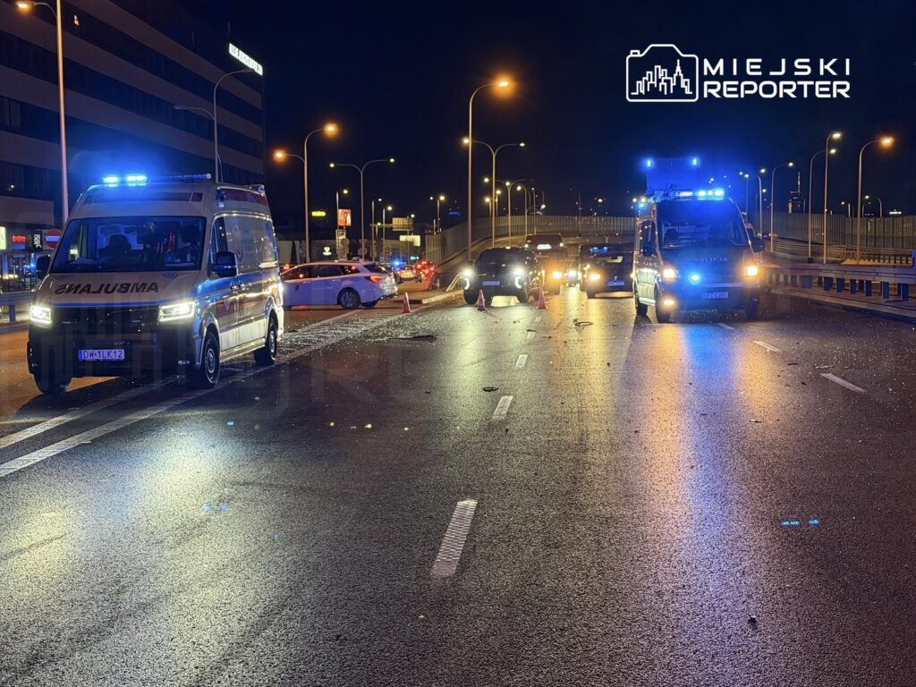 Ambulans i radiowóz policyjny na zablokowanej drodze, otoczone przez pojazdy i trójkąty ostrzegawcze w nocy.