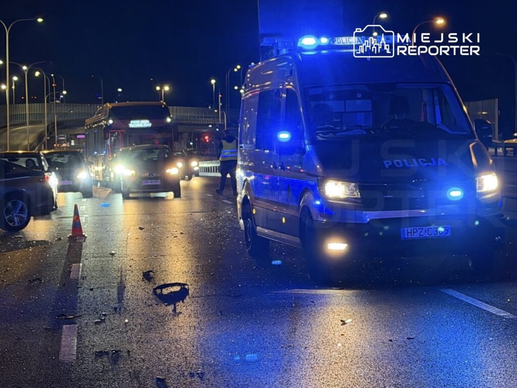 Na drodze w nocy, samochód policyjny z włączonymi sygnałami świetlnymi zabezpiecza miejsce wypadku, obok leżą resztki szkła.