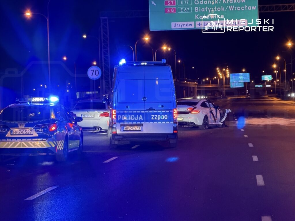 Na drodze nocą stoją dwa radiowozy policyjne obok uszkodzonego białego samochodu osobowego.