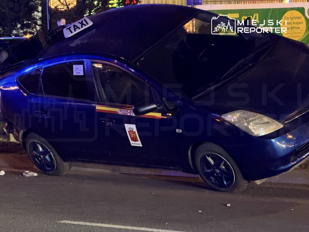 Zniszczony samochód osobowy z napisem "TAXI" na dachu, leżący na boku na ulicy w nocy.