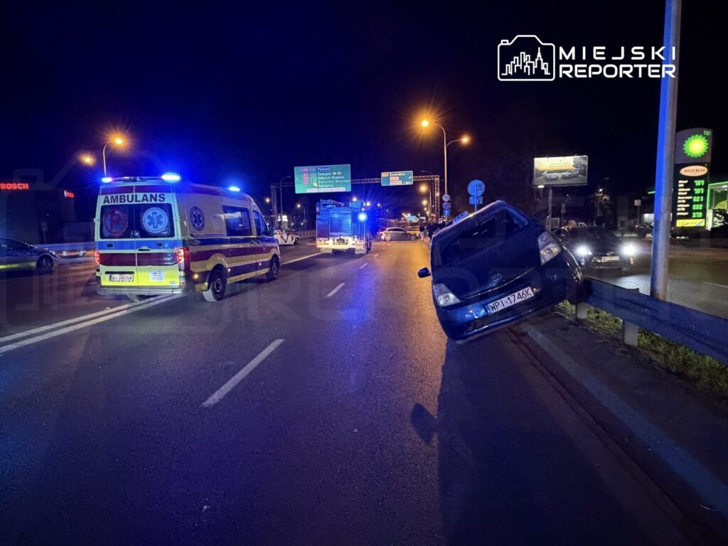 Samochód osobowy zawieszony na barierce drogowej obok karetki pogotowia i radiowozu w nocy na ruchliwej ulicy.