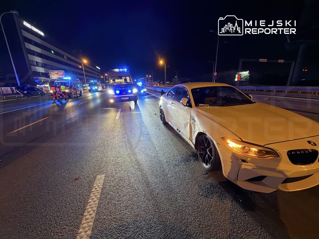 Biały samochód osobowy z uszkodzeniami stoi na drodze, obok interweniują pojazdy służb ratunkowych i policyjnych.