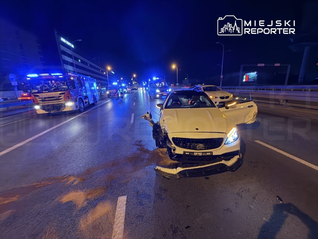 W nocy na drodze stoi uszkodzony biały samochód osobowy, obok którego widać pojazdy ratunkowe z włączonymi sygnałami świetlnymi.