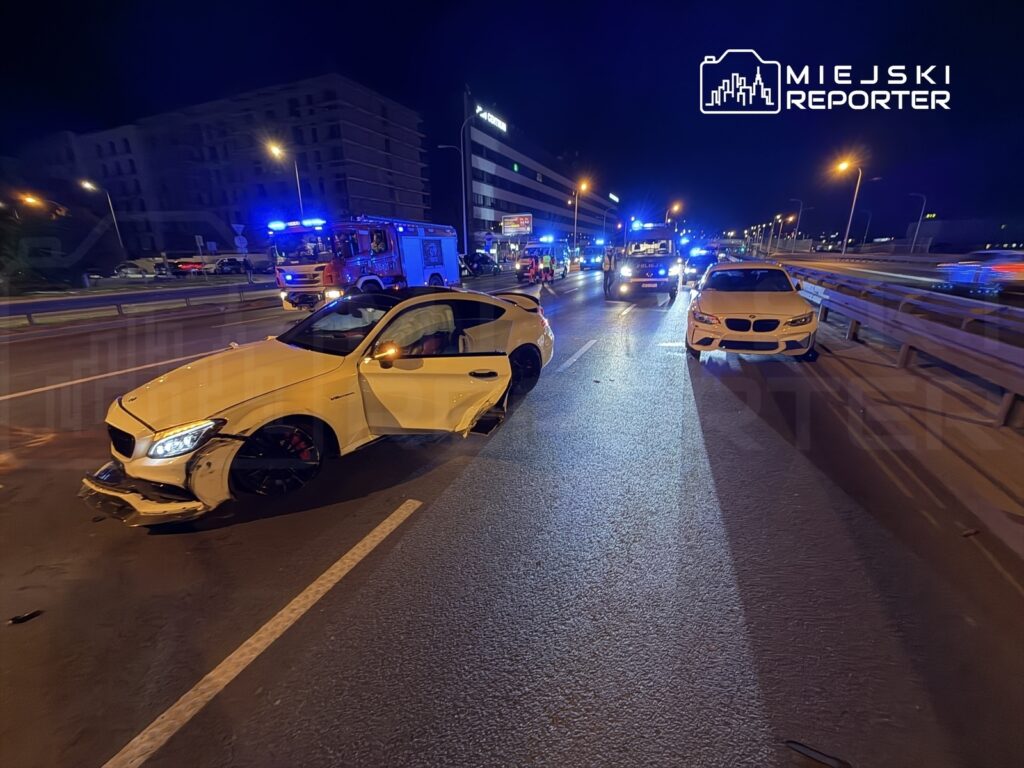 Uszkodzony biały samochód osobowy z otwartymi drzwiami stoi na drodze, obok widać niebieskie światła pojazdów ratunkowych.