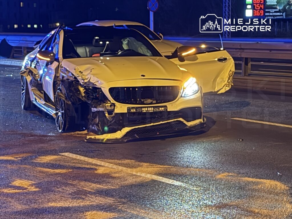 Uszkodzony biały samochód osobowy z wgniecioną karoserią stoi na jezdni w nocy, obok widoczne znaki drogowe.