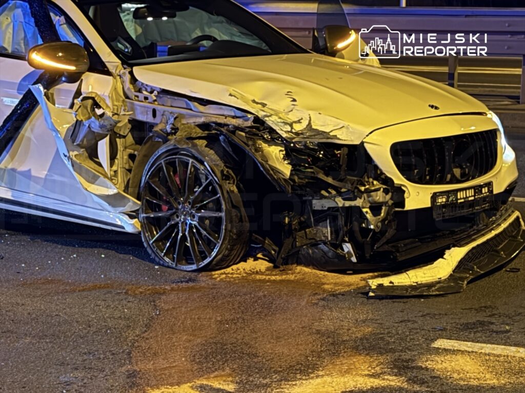 Uszkodzony biały samochód osobowy z poważnymi wgnieceniami z przodu i po stronie kierowcy na drodze nocą.