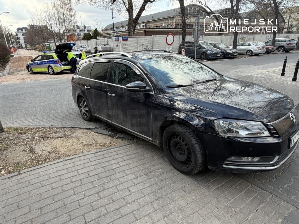 Czarny samochód osobowy zaparkowany na krawężniku obok radiowozu policyjnego, w tle widoczni funkcjonariusze.
