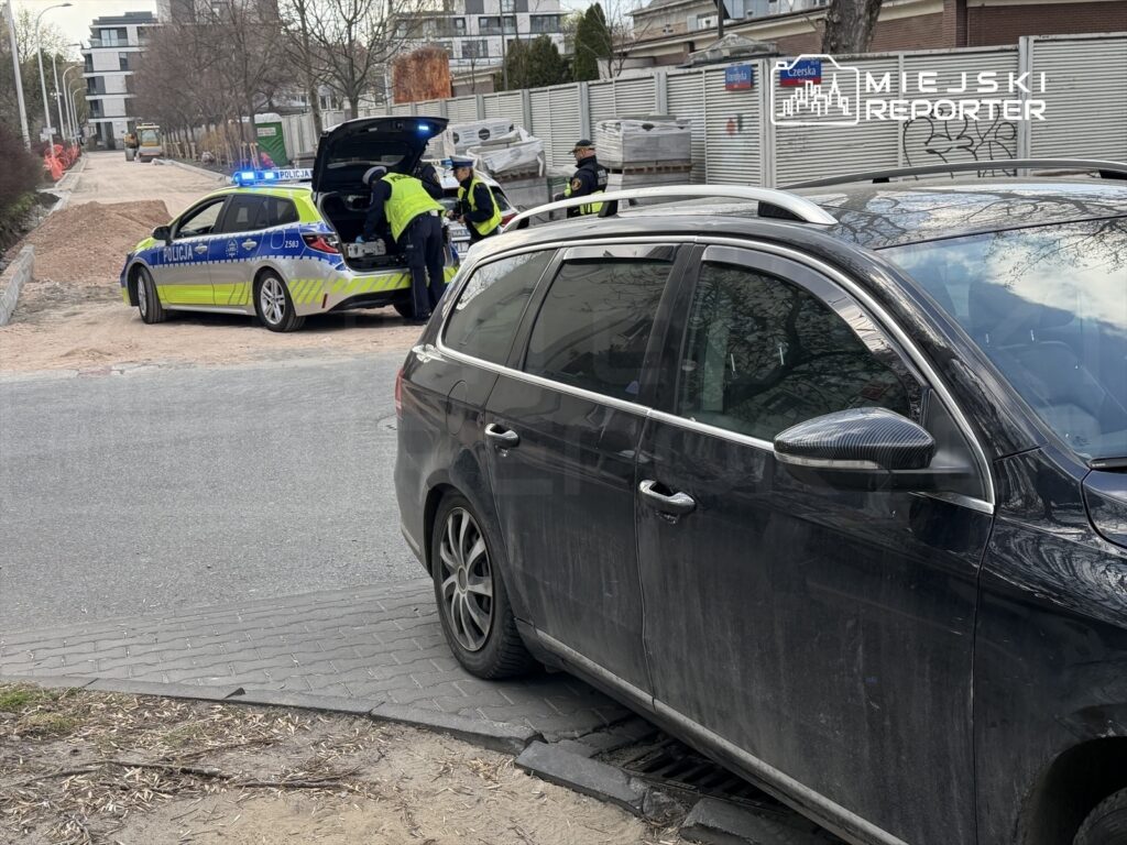 Policjanci przeszukują bagażnik radiowozu na tle drogi w trakcie remontu, obok stoi czarny samochód osobowy.