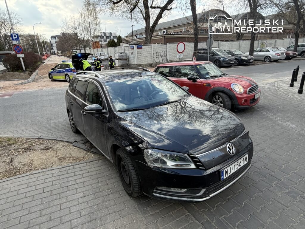 Czarny samochód osobowy zaparkowany na chodniku, obok policjanci rozmawiający przy radiowozie.