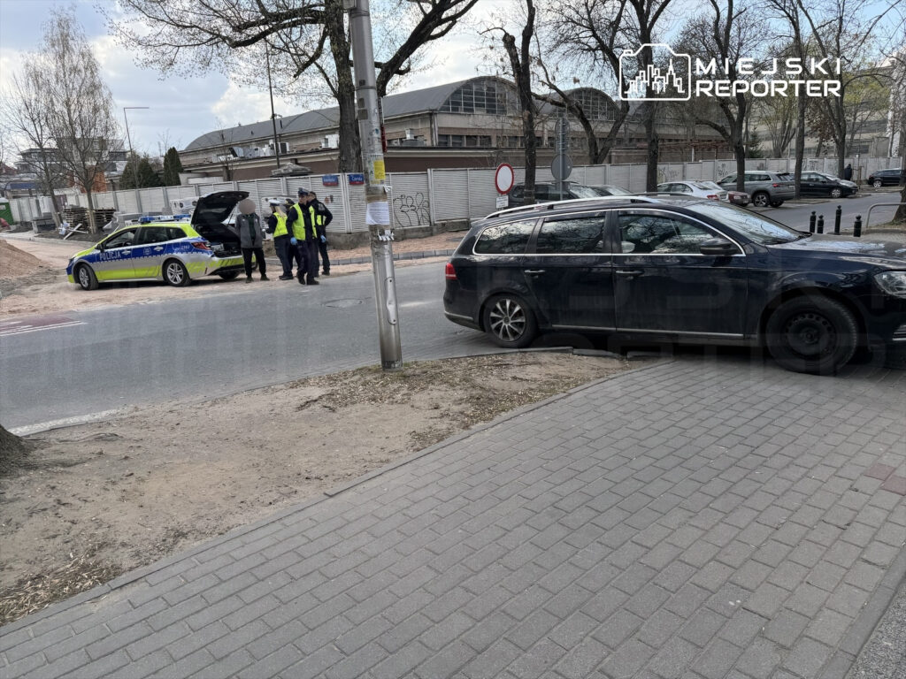 Grupa policjantów w odblaskowych kamizelkach rozmawia z mężczyzną obok radiowozu na ulicy.