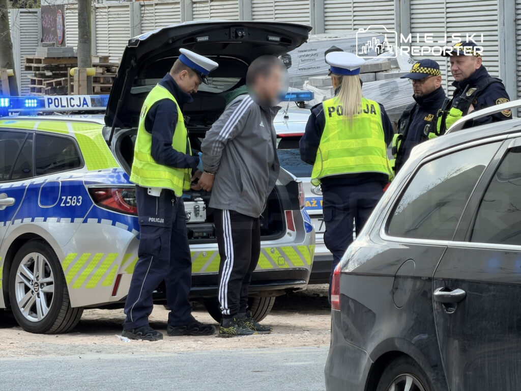 Mężczyzna w szarej dresowej odzieży jest zatrzymywany przez policjantów w kamizelkach odblaskowych obok radiowozu.