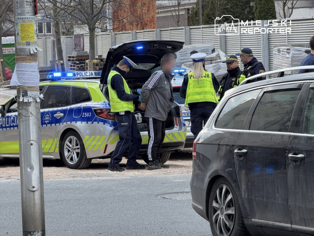 Mężczyzna w szarej bluzie jest zatrzymywany przez policjantów obok radiowozu na miejskiej ulicy.