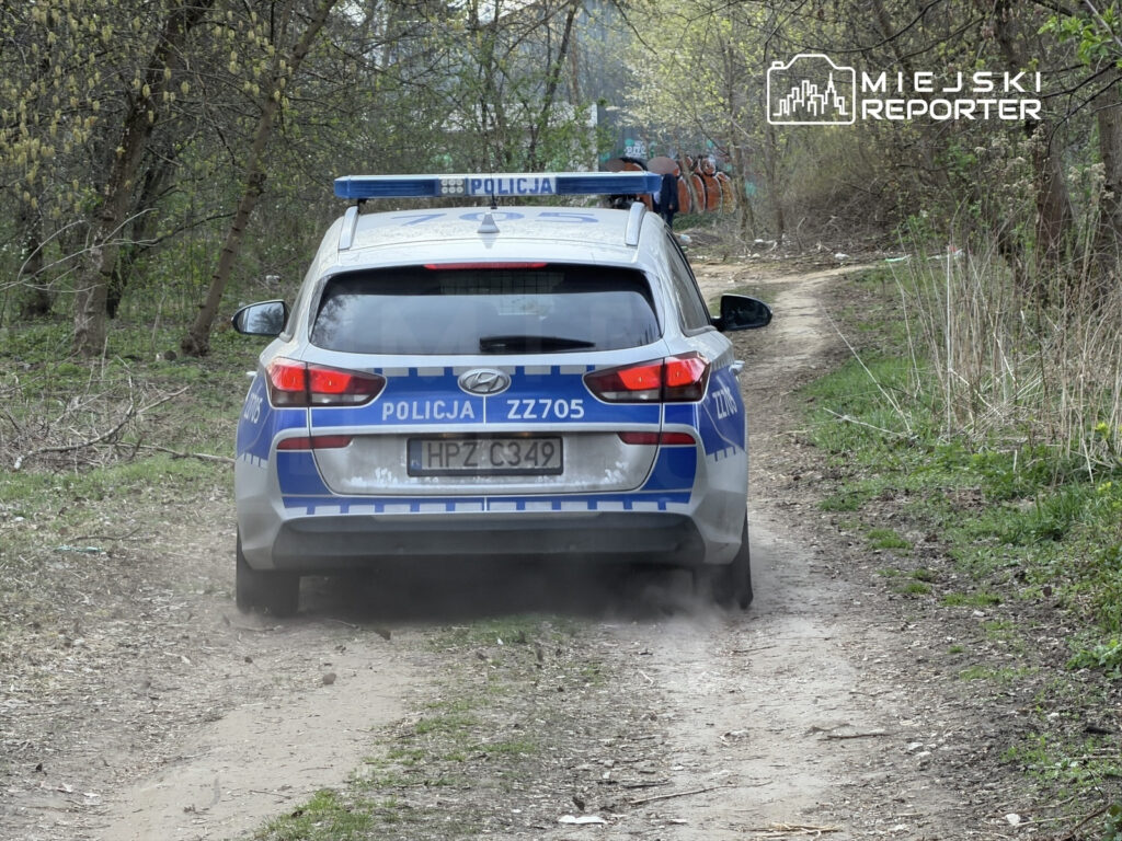 Radiowóz policyjny porusza się po wąskiej, wyboistej drodze w otoczeniu drzew i krzewów.