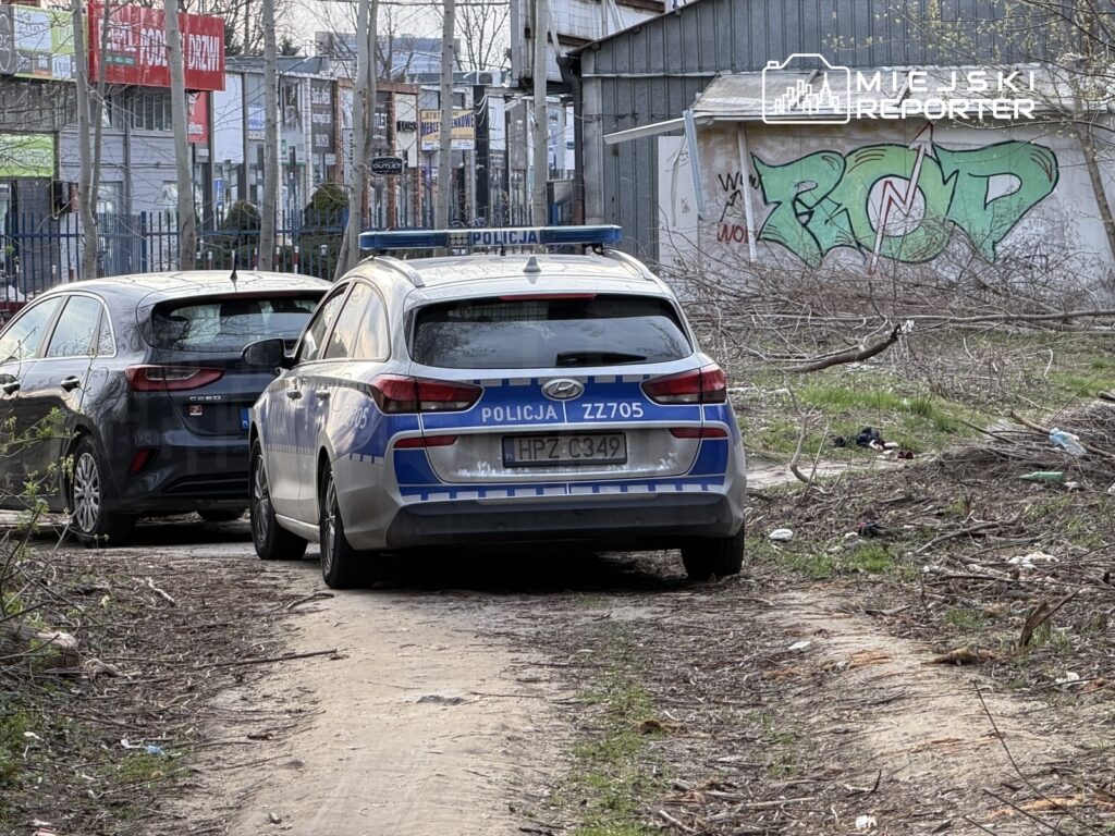 Policyjny radiowóz zaparkowany na nieutwardzonej drodze obok ciemnego samochodu, w tle widoczna graffiti na ścianie.