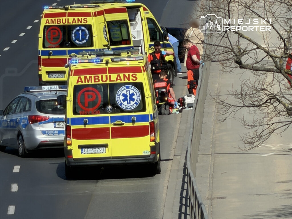 Zespół ratunkowy w mundurach udziela pomocy na poboczu drogi, obok dwóch karetek i radiowozu policji.