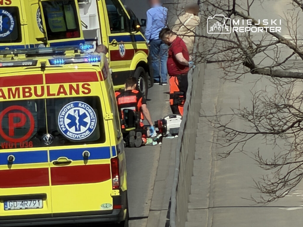 Ratownicy medyczni udzielają pomocy na chodniku obok karetki, w otoczeniu zarośli i przechodniów.