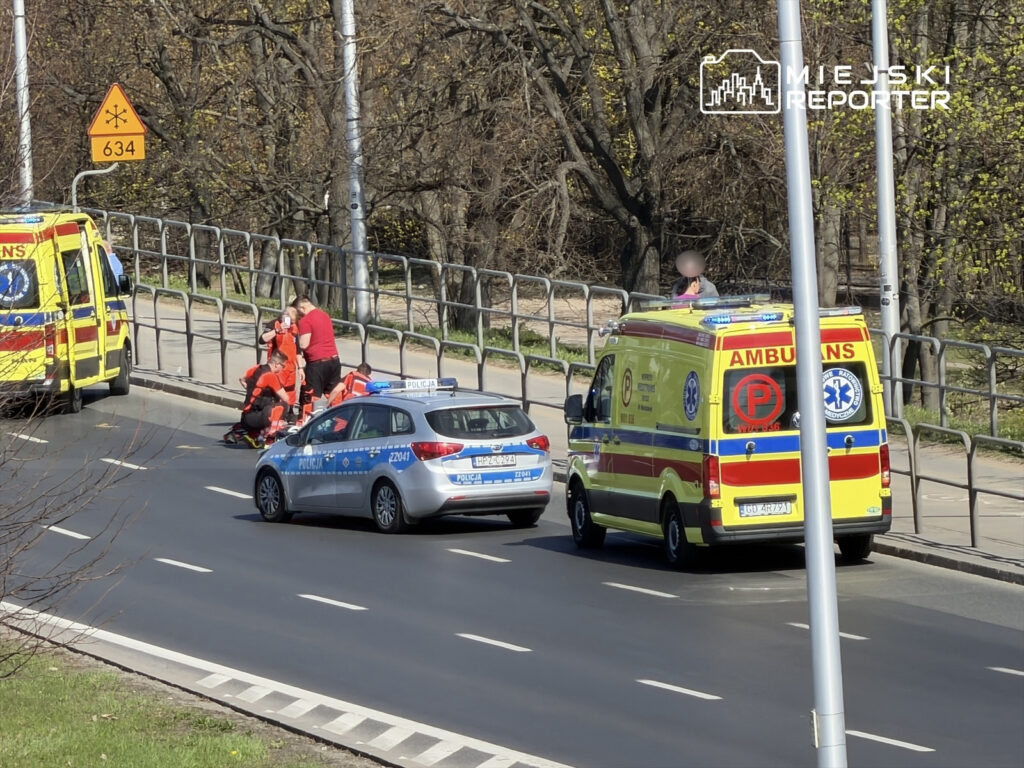 Zespół ratowników medycznych udziela pomocy osobie poszkodowanej na chodniku, obok policyjnego radiowozu i karetki.