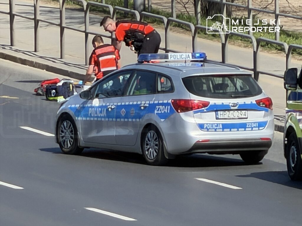 Dwaj ratownicy w pomarańczowych kamizelkach udzielają pomocy osobie leżącej na jezdni obok radiowozu policyjnego.