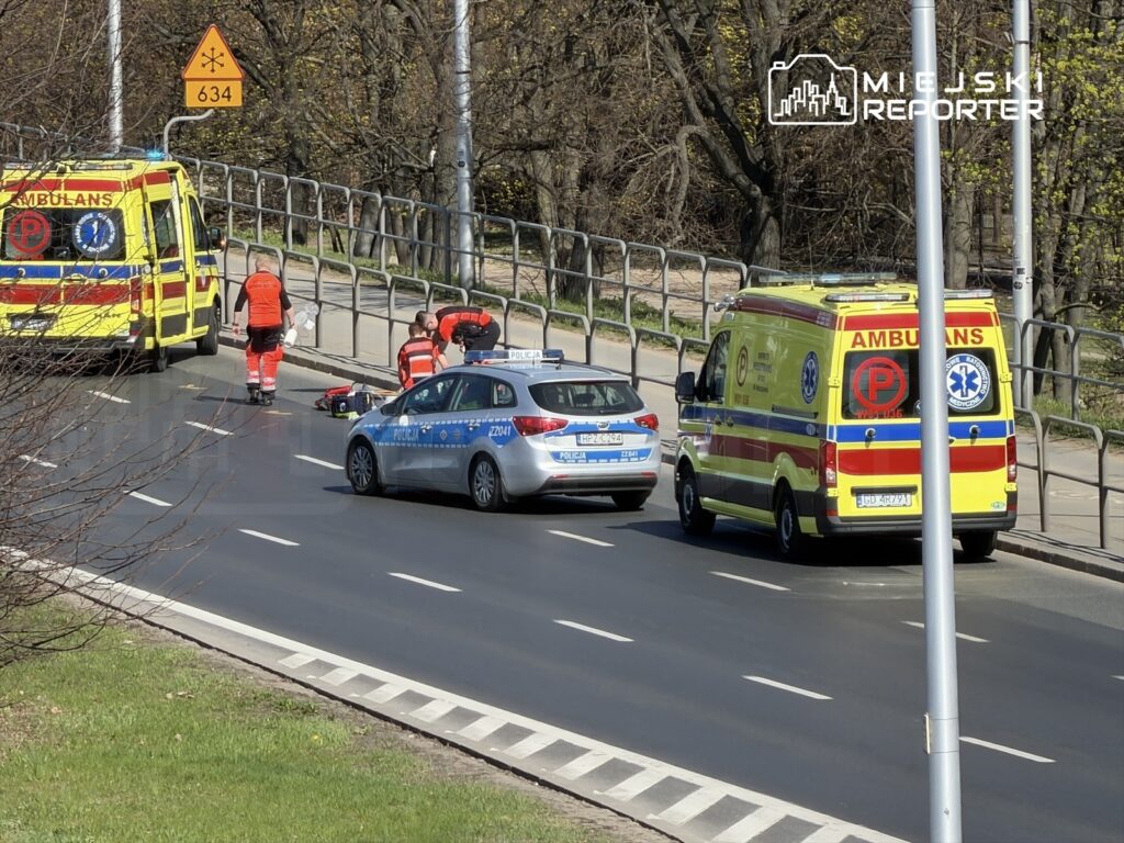 Ratownicy medyczni udzielają pomocy poszkodowanemu na jezdni, obok policyjnego samochodu i karetki.