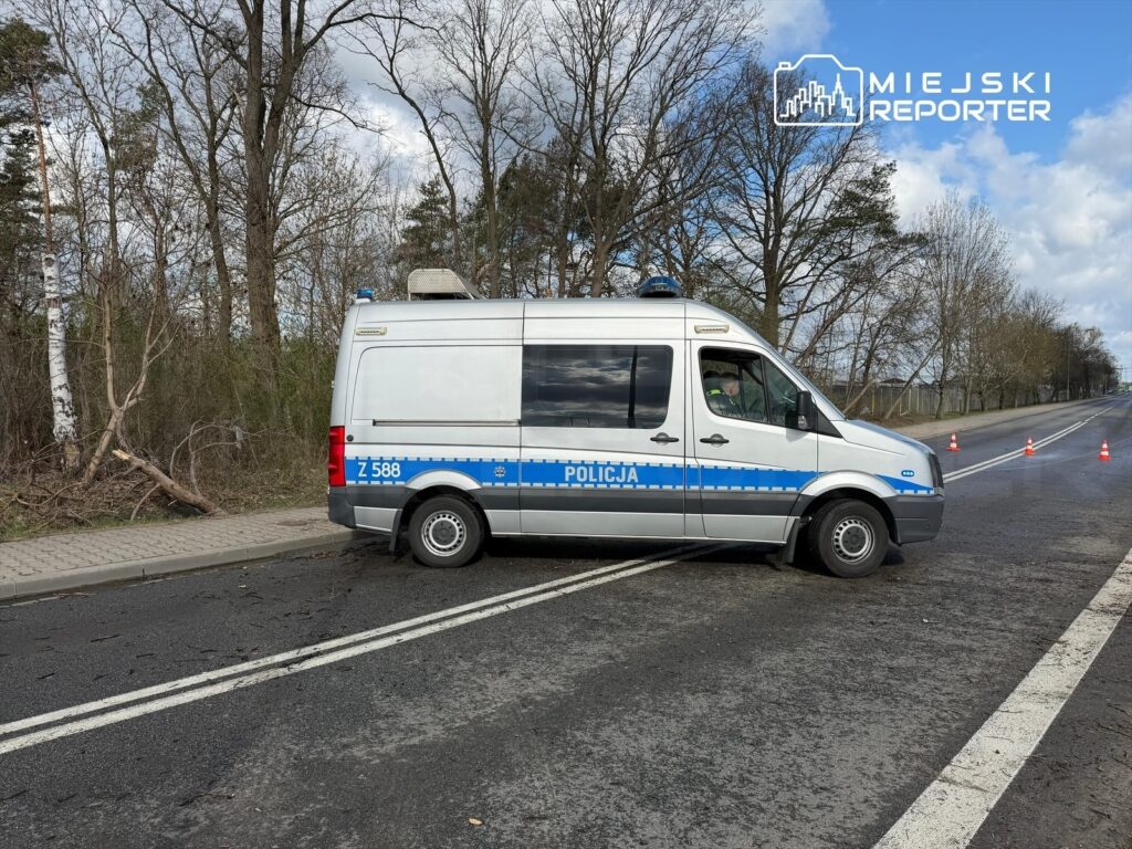 Policyjny furgon z niebieskimi oznaczeniami stoi na drodze, obok znaków ostrzegawczych i drzew w tle.