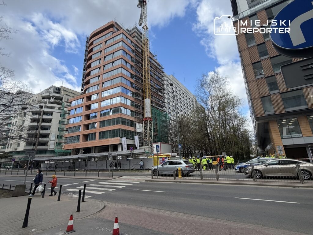 Grupa pracowników budowlanych w kamizelkach odblaskowych prowadzi prace przy nowoczesnym biurowcu z przeszkloną fasadą.