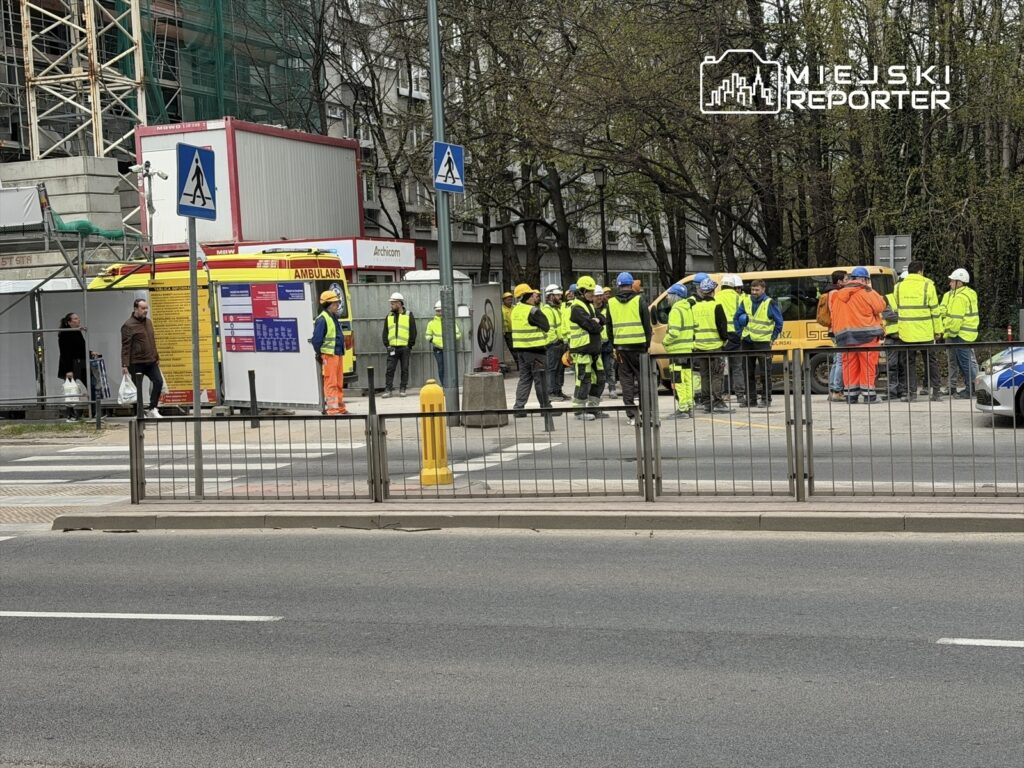 Grupa pracowników budowlanych w odblaskowych kamizelkach zbiera się w pobliżu placu budowy, obok ambulansu.