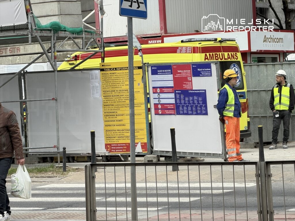 Mężczyzna w brązowej kurtce przechodzi obok budowy, na której pracownicy w kamizelkach odblaskowych rozmawiają przy ambulansie.