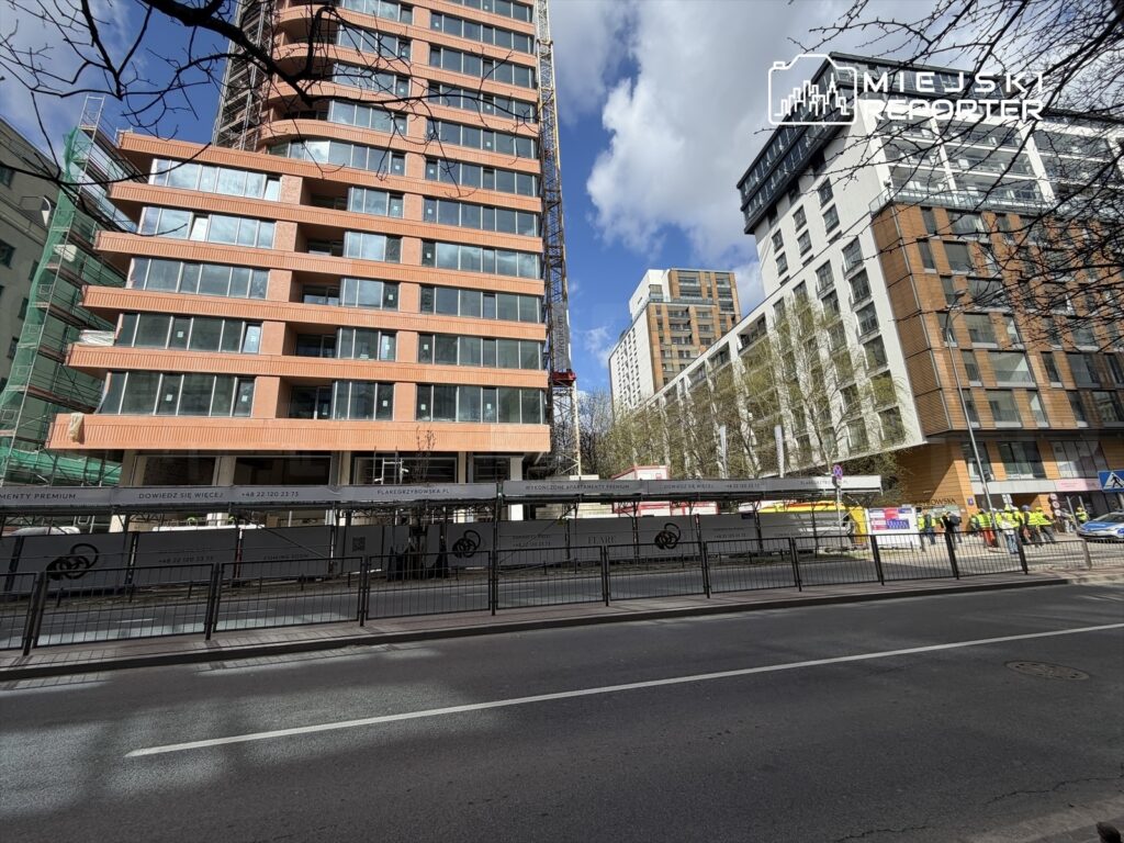 Nowoczesny budynek mieszkalny w trakcie budowy, z widocznymi oknami i rusztowaniami, otoczony przez pracowników na ulicy.