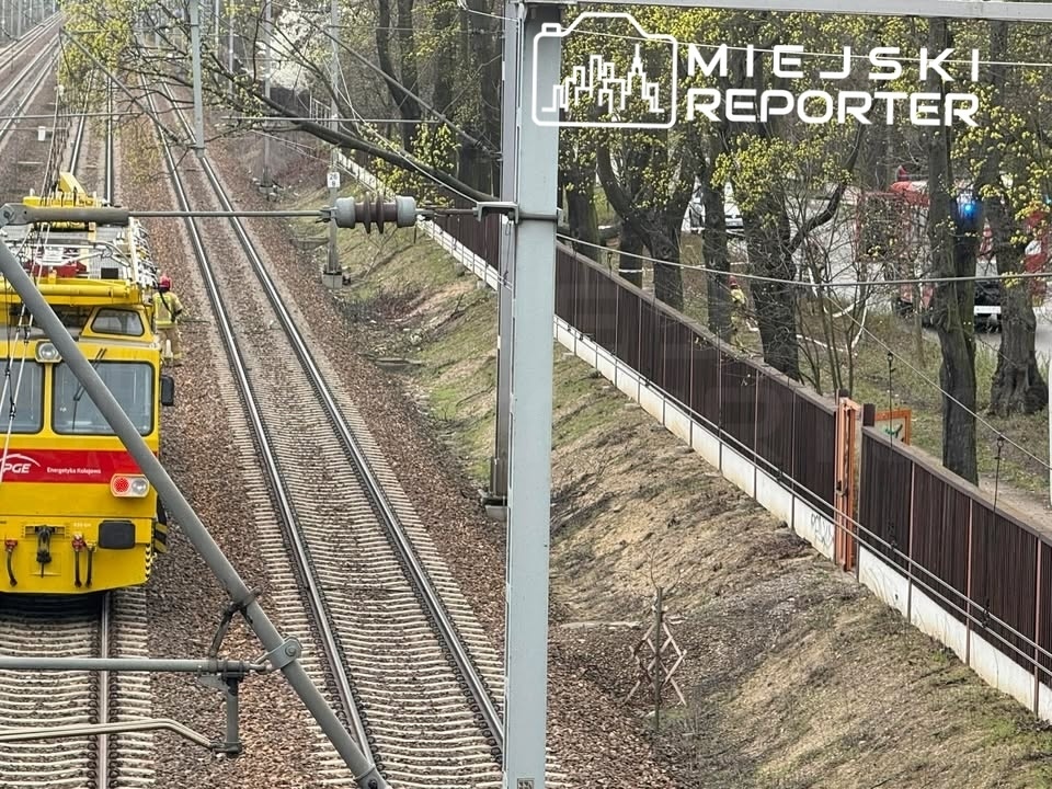 Żółty pojazd kolejowy na torach, z drzewami w tle oraz ogrodzeniem wzdłuż linii kolejowej.