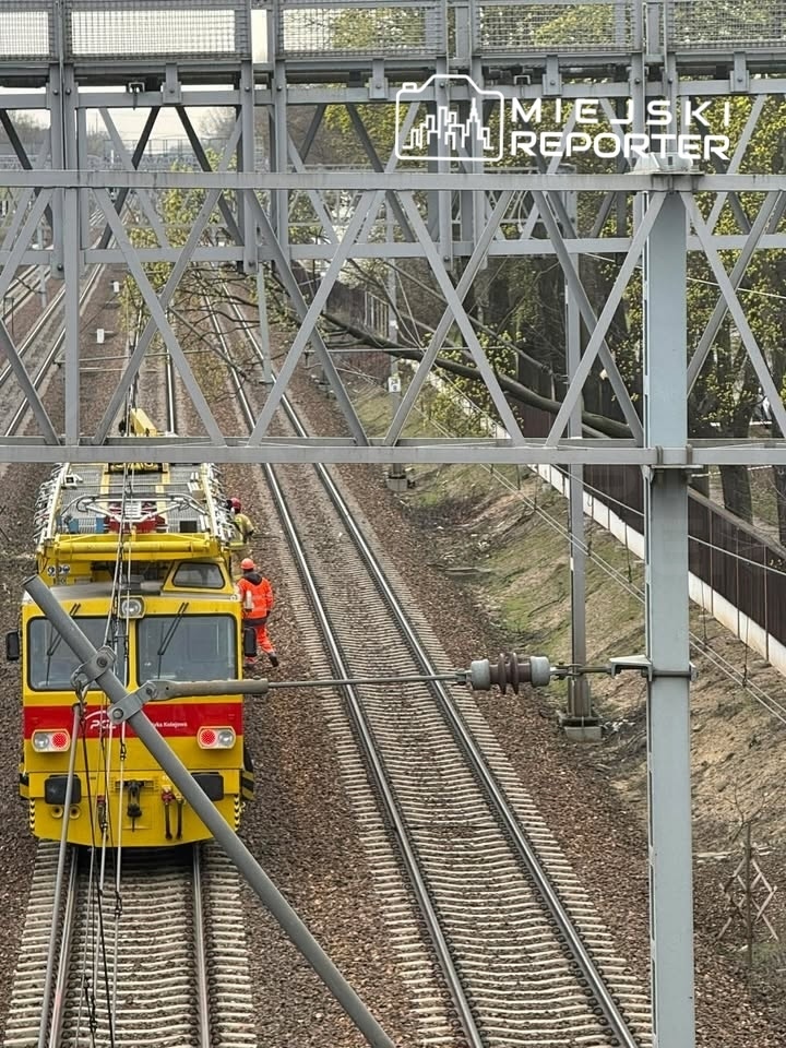 Pojazd kolejowy w żółto-czerwonym malowaniu zatrzymany na torach, pracownicy w pomarańczowych kamizelkach dokonują inspekcji.