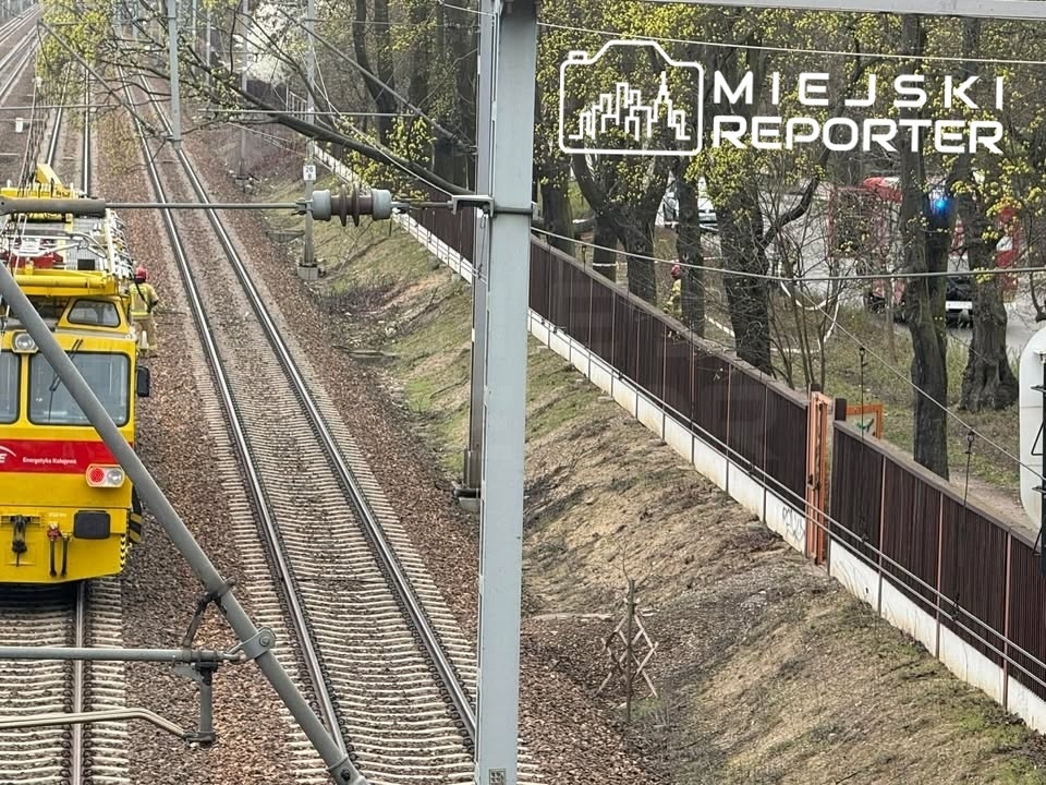 Żółty pociąg na torach, obok widoczne drzewa i ogrodzenie, w tle fragment drogi.