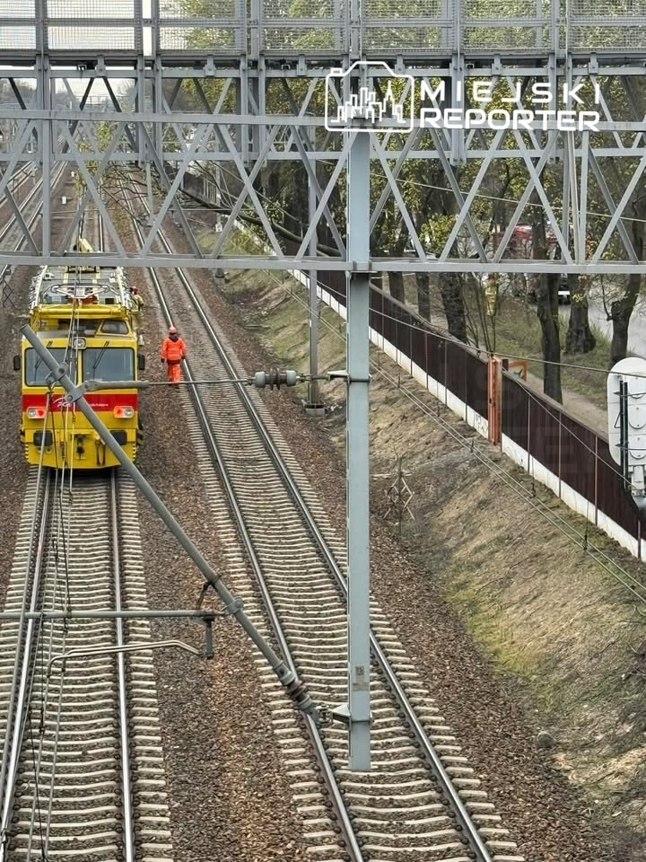 Robotnik w pomarańczowym uniformie pracuje przy torach kolejowych obok żółtej lokomotywy.