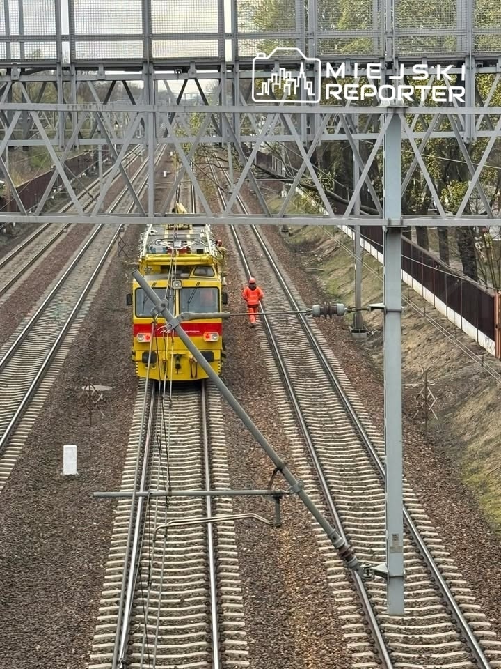 Pracownik w pomarańczowym kombinezonie kieruje ruchem obok żółtej lokomotywy na torach kolejowych.