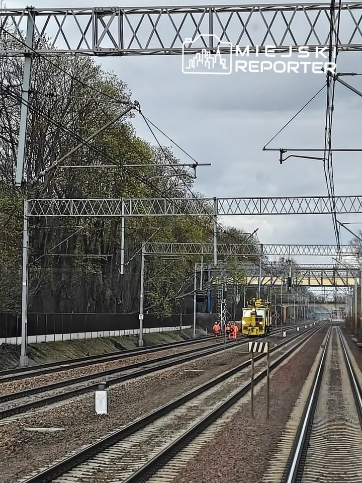 Pracownicy w pomarańczowych kamizelkach dokonują napraw na torach kolejowych obok żółtej maszyny roboczej.