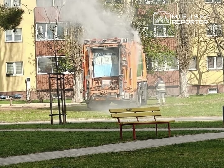 Śmieciarka wydobywająca dym, stojąca na trawniku przed blokiem mieszkalnym, obok pracownik w odzieży roboczej.