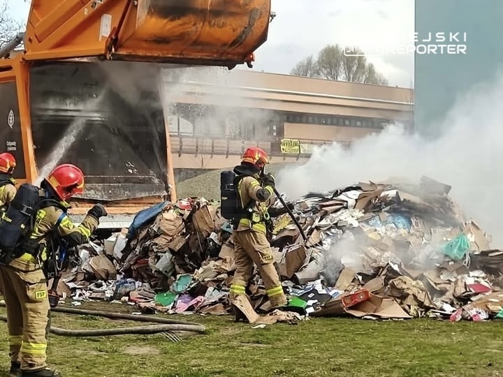 Strażacy gaszą pożar odpadów w kontenerze, z którego wydobywa się dym, w miejskim otoczeniu.