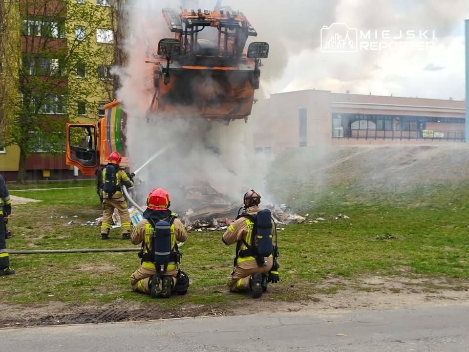 Strażacy gaszą pożar, który wybuchł przy wysypisku, obok podnoszącego się śmieciarki w tle. W powietrzu unosi się dym.