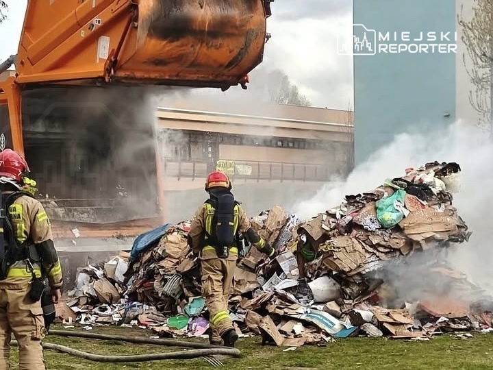 Strażacy w hełmach gaszą palący się stos odpadów, podczas gdy śmieciarka podnosi kontener. W tle widoczny budynek.