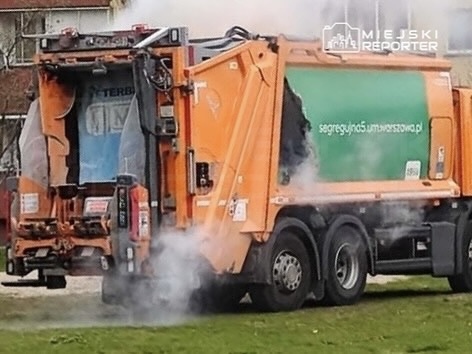 Śmieciarka z uniesioną klapą, wydobywająca dym, stoi na trawniku obok budynków mieszkalnych.