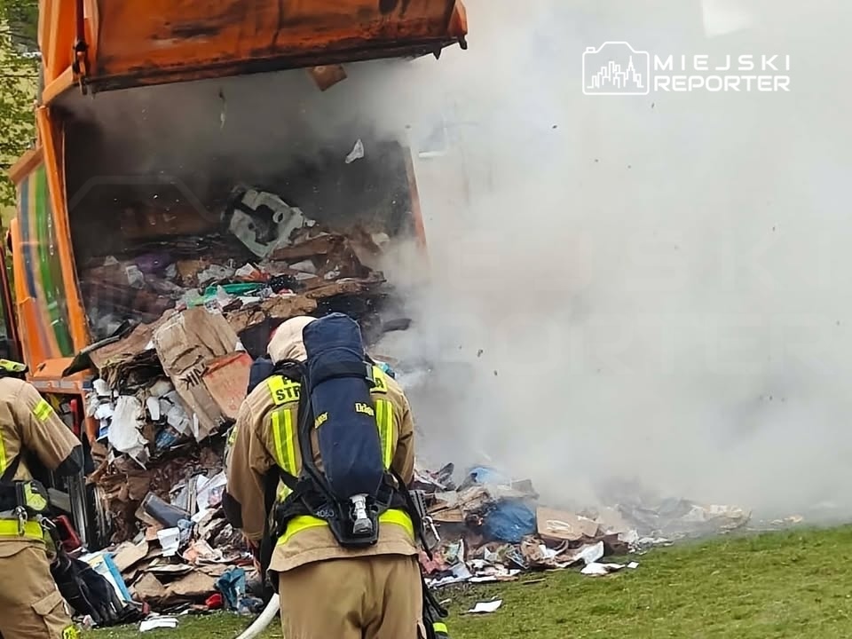 Strażacy z wężami gaśniczymi gaszą pożar wydobywający się z kontenera na odpady. W tle widoczny dym.