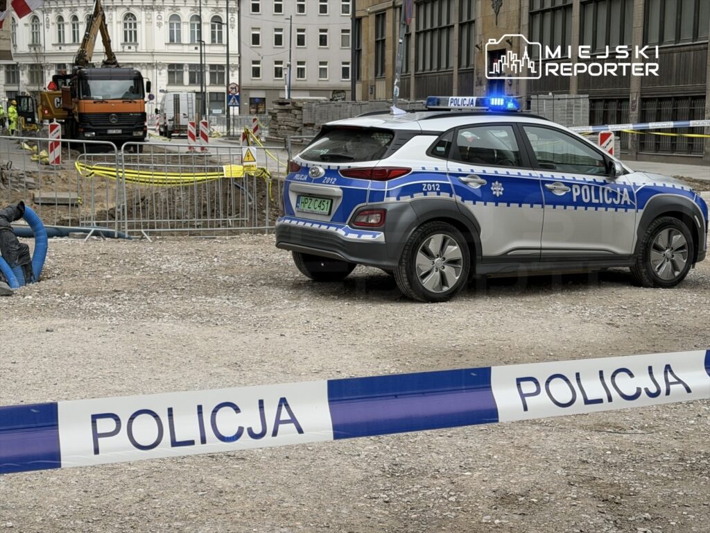 Policyjny samochód osobowy zaparkowany przy ogrodzeniu budowy, obok pracowników w odzieży roboczej.