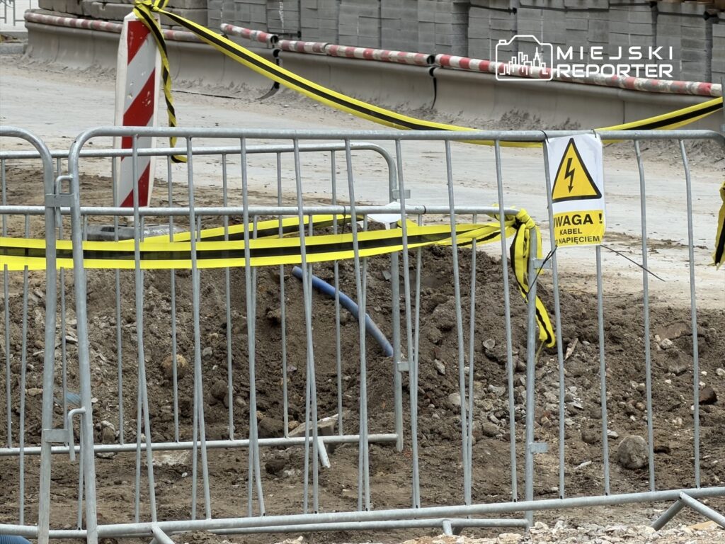 Ogrodzenie zabezpieczające wokół wykopu, oznaczone ostrzeżeniem o niebezpieczeństwie elektrycznym.