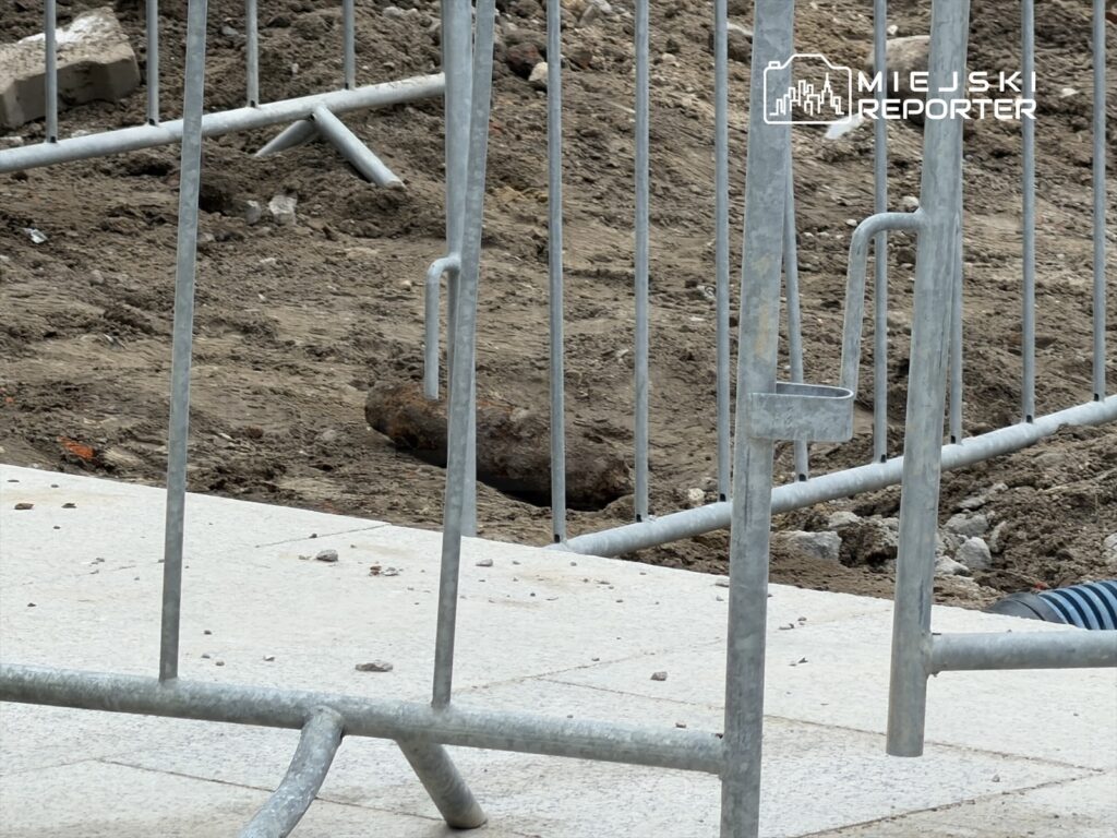 Robotnicy zabezpieczają teren budowy ogrodzeniem, na którym leży nieokreślony obiekt w ziemi.