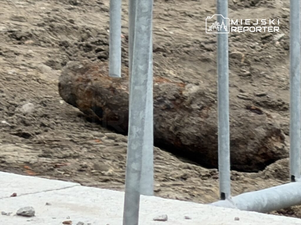 Rdzewiejący pocisk leży na odkrytej ziemi, otoczony metalowym ogrodzeniem.