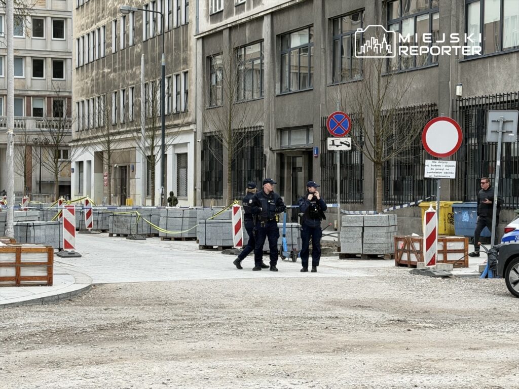 Trzech policjantów w mundurach patroluje zablokowaną ulicę, wokół znajdują się materiały budowlane i oznakowania.
