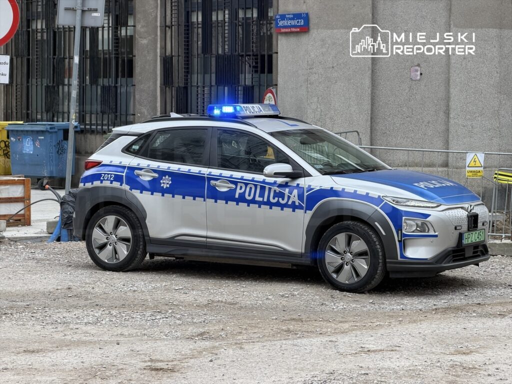 Policyjny samochód osobowy z niebieskimi światłami zaparkowany na placu budowy w miejskim otoczeniu.